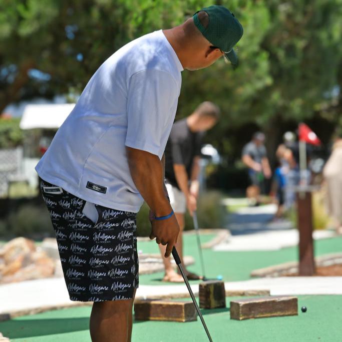 A golfer is captured mid-putt, showcasing the Legacy Shorts from KLMulliganz. These black shorts, featuring a bold, all-over print of the “KLMulliganz” logo, combine style and performance, perfect for both the golf course and everyday wear. The Legacy Shorts are highlighted as the focal point of the image, representing the brand’s commitment to quality golf apparel that seamlessly transitions into a lifestyle statement. The golfer’s stance, paired with the vibrant outdoor setting, underscores the versatilit